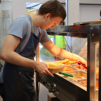 Person serving hot food into a takeaway container at a cafeteria counter.